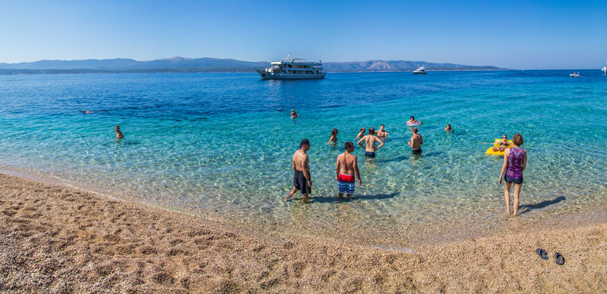 A group of beachgoers swimming from a sandy beach in blue water with a small ship floating in the distance. 