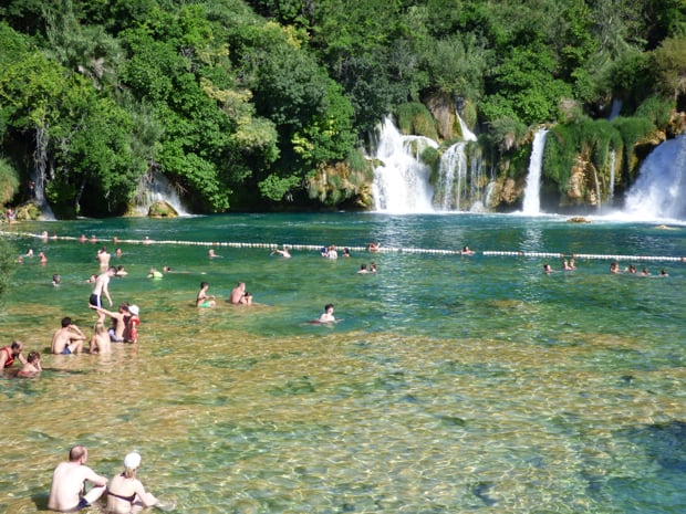 Krka National Park in Croatia with several waterfalls dropping into a big swimming hole with several tourists swimming or sitting about.