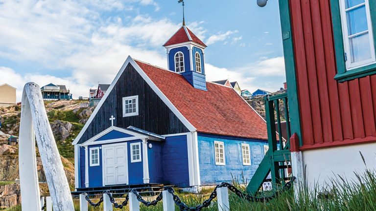 Town of Sisimut, Greenland, on a clear day, with light blue church with red roof, steeple & white trim, beside a red & green building.