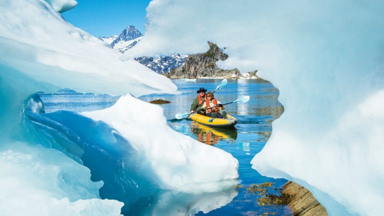 Tandem kayakers paddle around icebergs in glassy water on a sunny day near Skjoldungen Island, Southeastern Greenland.
