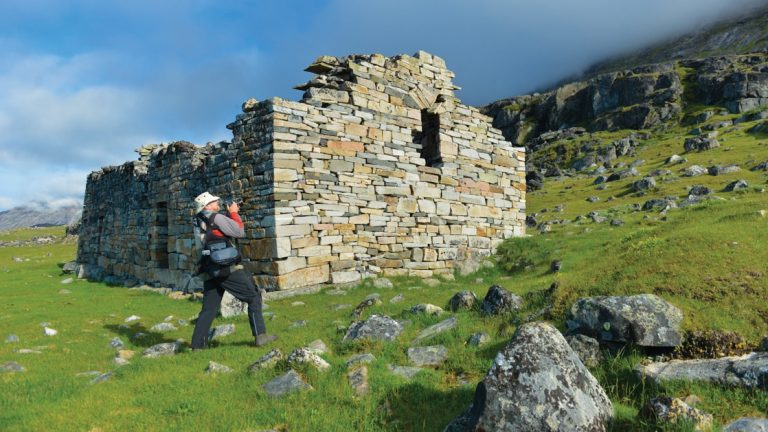 Traveler visits ruins of a stone hut set on a grassy hillside, seen on the Iceland's Wild West Coast to East Greenland Cruise.