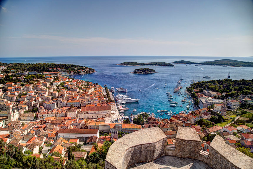 Aerial view of a Croatian port town with terra cotta roofs, beige stone buildings, lush green hillsides, docked small ships & turquoise water.