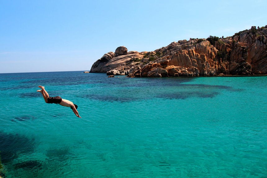 Man dives into turquoise water beside red rocky shoreline on a Mediterranean small ship cruise.