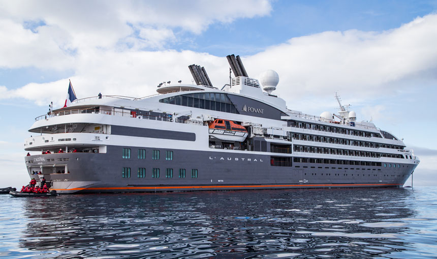 Large gray & white expedition ship with French flag sits in glassy water as travelers disembark onto black Zodiacs.