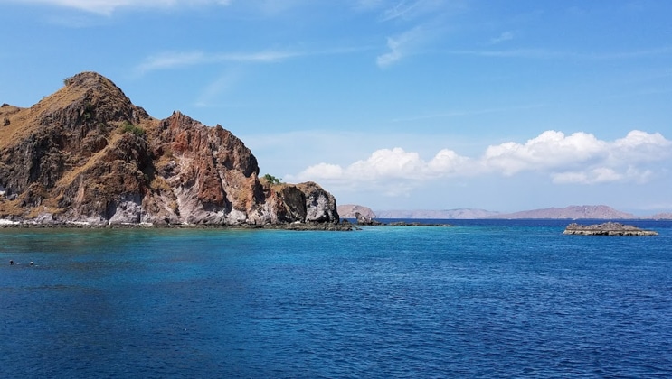 Bright blue & turquoise sea is calm beside a tan rocky shoreline on a sunny day during an cruise from Singapore to Bali.