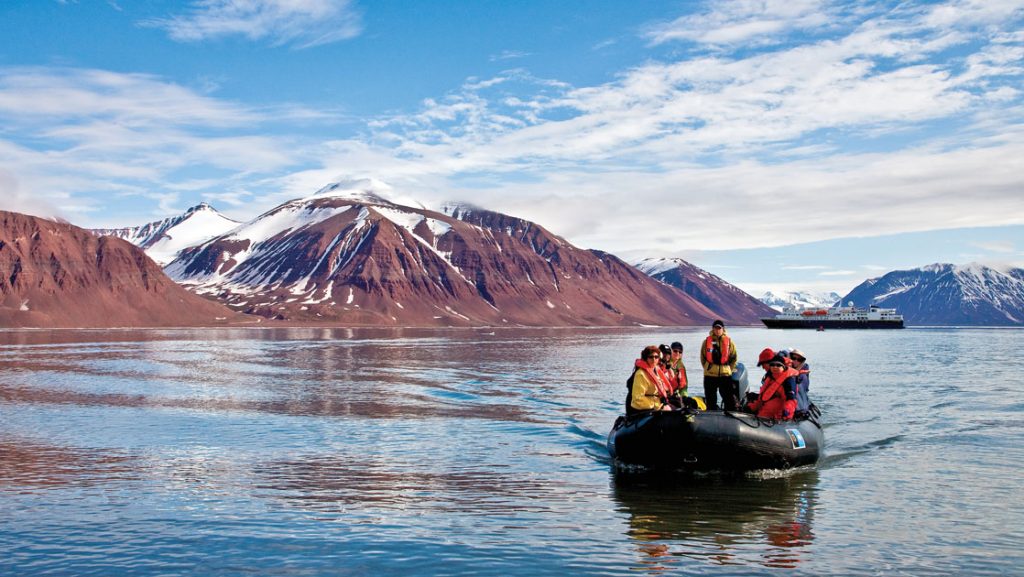 Polar travelers in a black Zodiac inflatable boat cruise in calm waters with brown & white mountains in the background.