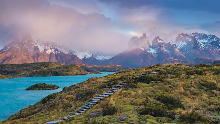 Aerial view of bright blue water beside snowcapped peaks & grassy hillside on a Patagonia to Antarctica cruise.