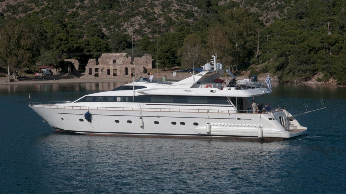 Aerial exterior view of white mega yacht at anchor beside ancient ruins & green hillsides in Greece.