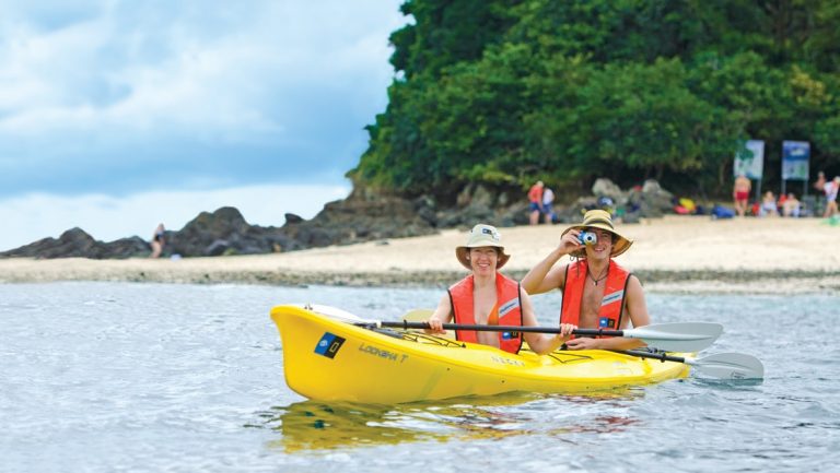 Two male travelers wear orange life jackets and tan wide brimmed hats paddle a yellow double kayak off the coast of Costa Rica.