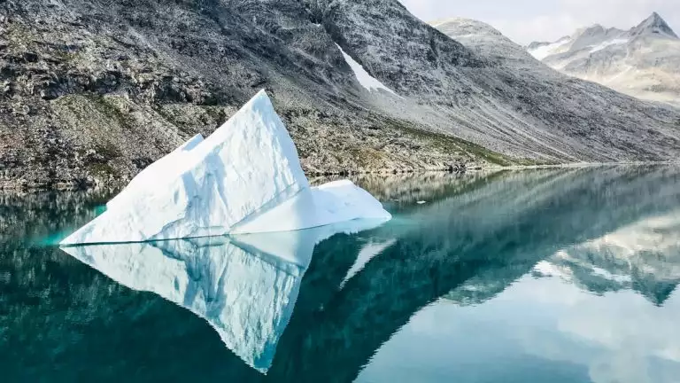 Tiny iceberg floats in glassy reflective waters of a fjord with rocky gray peaks beside, seen on the Wild Greenland Escape voyage.
