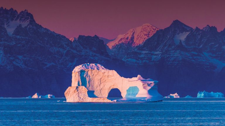 Large white iceberg with an arch in its center sits in purple light at dusk during the Wild Greenland Escape small ship voyage.
