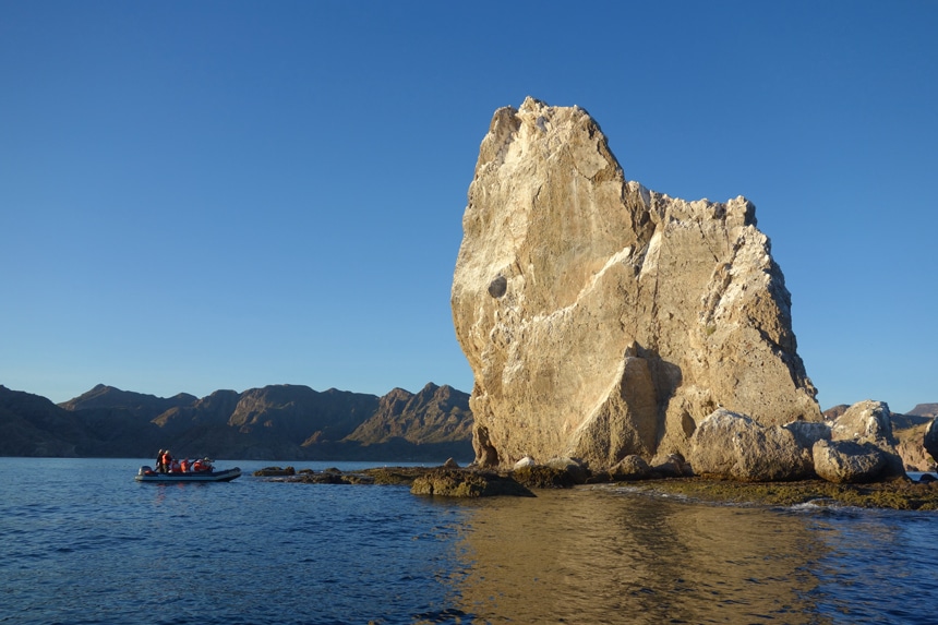 A large tan jagged rock formation extends from the ocean off the coast of Baja, a zodiac boat with passengers cruises nearby.
