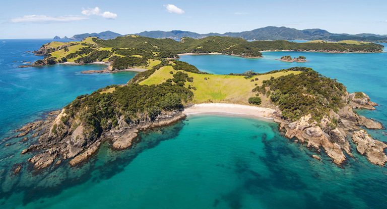 Aerial view of a long, skinny island with grassy and forested areas & white-sand beach area in New Zealand.