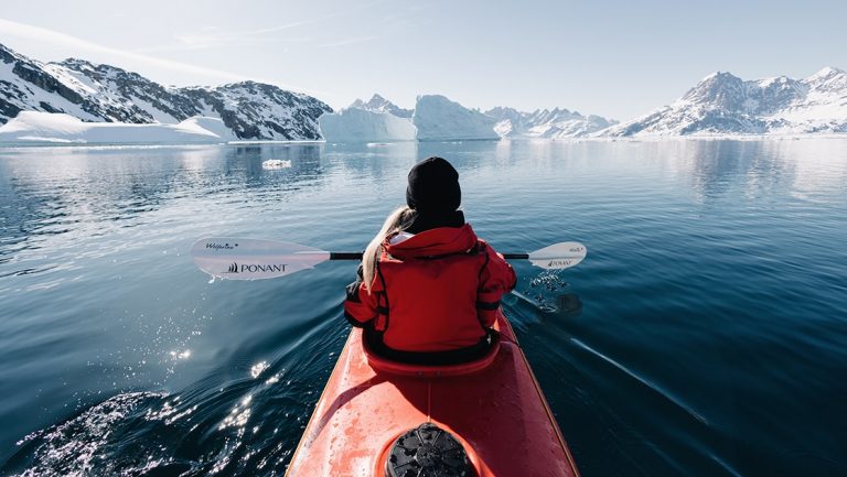 Female in red parka paddles red kayak through calm Arctic ocean surrounded by snow covered jagged mountains in Greenland.