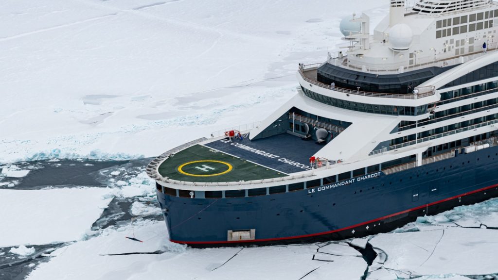 Aerial view of modern expedition ship with dark blue hull, white upper decks & helicopter pad on its bow, parked in ice at the North Pole.