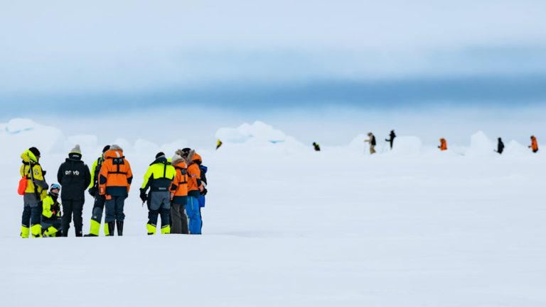 Group of polar travelers in bright jackets stand atop ice & snow during the Le Commandant Charcot North Pole Expedition.