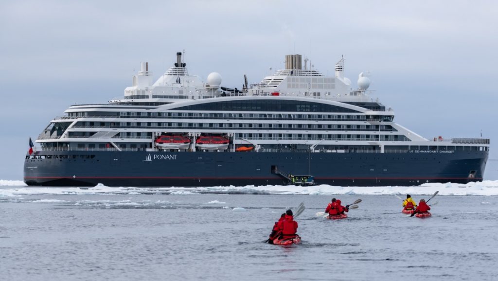 Dark blue & white expedition ship sits beside ice as travelers in red jackets & kayaks paddle beside on a luxury North Pole voyage.