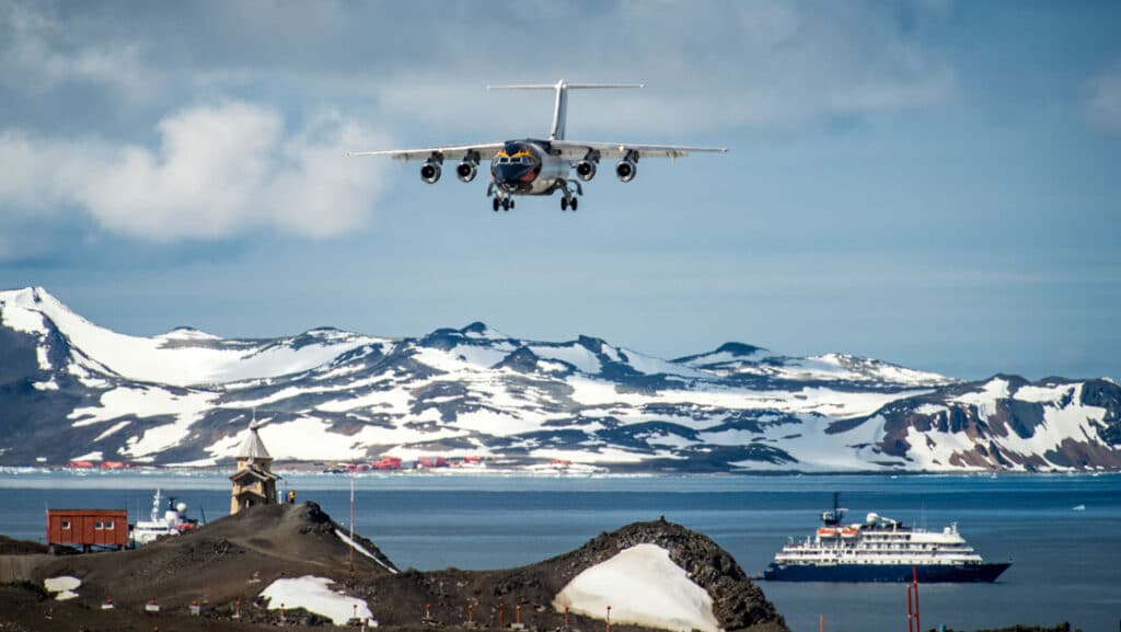 A plane seen landing on Antarctica at King George Island with snowy mountains and a ship out at anchor