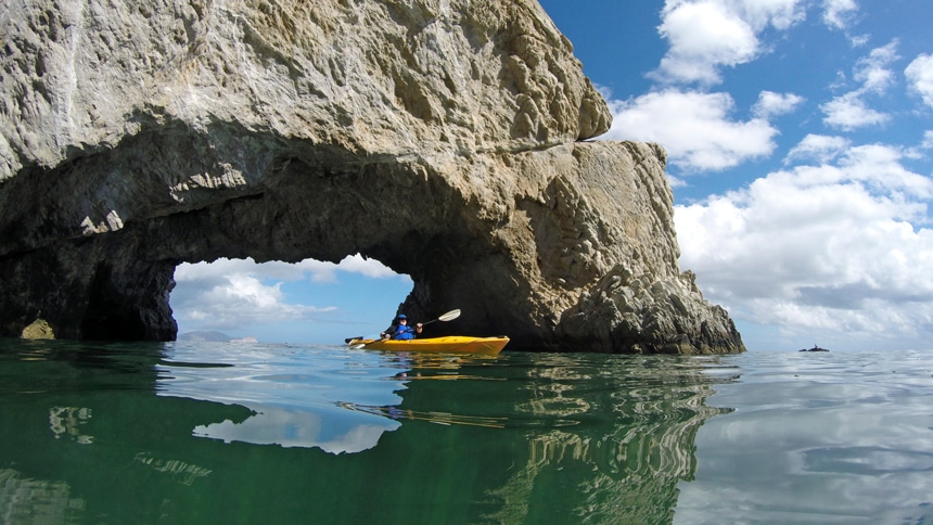 White clouds dot the bright blue sky as a man in a yellow kayak paddles through a rock arch formation in Baja.