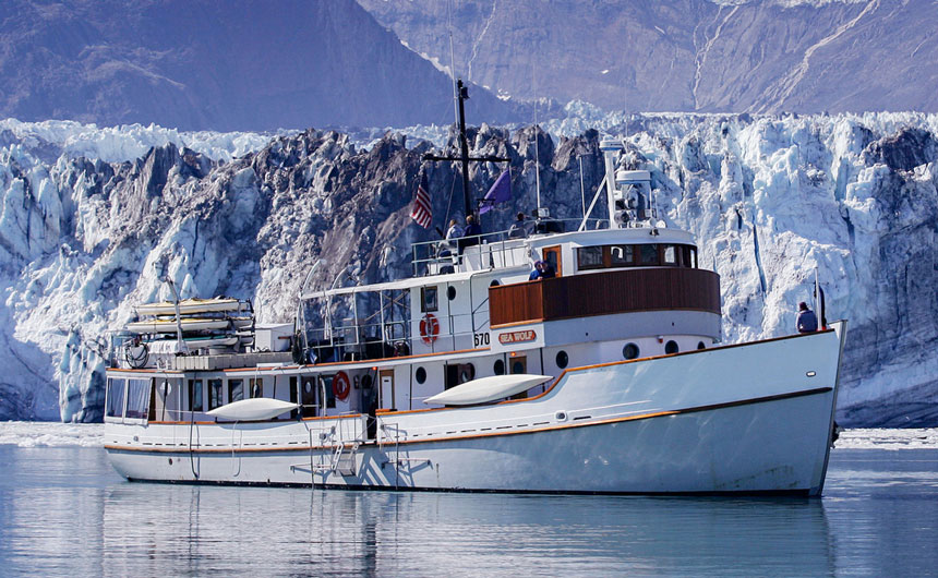 An Alaska small ship, the Sea Wolf, floats in front of a jagged white and grey glacier, beyond them, a sweeping purple hued Alaska mountain scape.