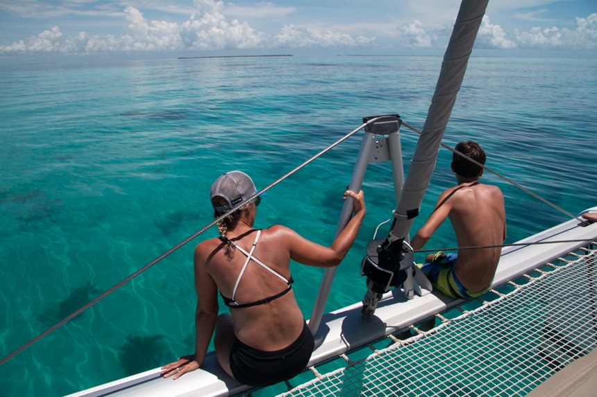 A male and female dressed in swim gear sit on the edge of a catamaran looking out over the teal blue ocean water at the horizon in Belize. 