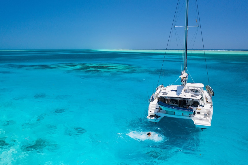 A boat guests swims around an all white catamaran has it floats in clear blue ocean water that matches the clear blue sky above it. 