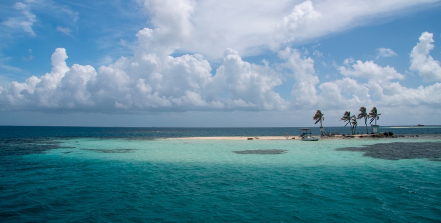 White clouds line the blue sky, hovering above a palm tree filled small sandy island surrounded by teal ocean in Belize. 