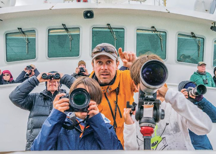At the bow of a boat, children use cameras and binoculars as they are instructed by a Lindblad Expeditions professional photographer. 