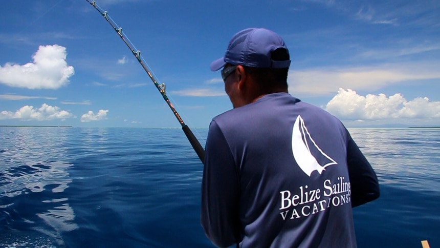 On a blue sky day in Belize, a man wearing a navy blue hat and long sleeve shirt hold a rod and fishes from the back of a boat. 