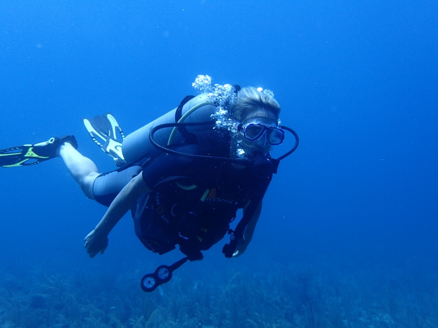 A deep blue underwater photo of a solo female scuba diver wearing fins, a mask, a snorkel, a wetsuit and other dive equipment