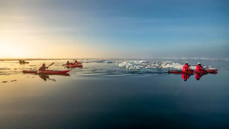 Tandem kayakers in red boats paddle glassy water among small icebergs at dusk on an Arctic cruise near the North Pole.