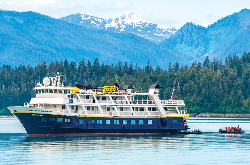 The blue and white boat, the sea bird, floats in Alaska in front of green forested shoreline and snow capped mountain range.
