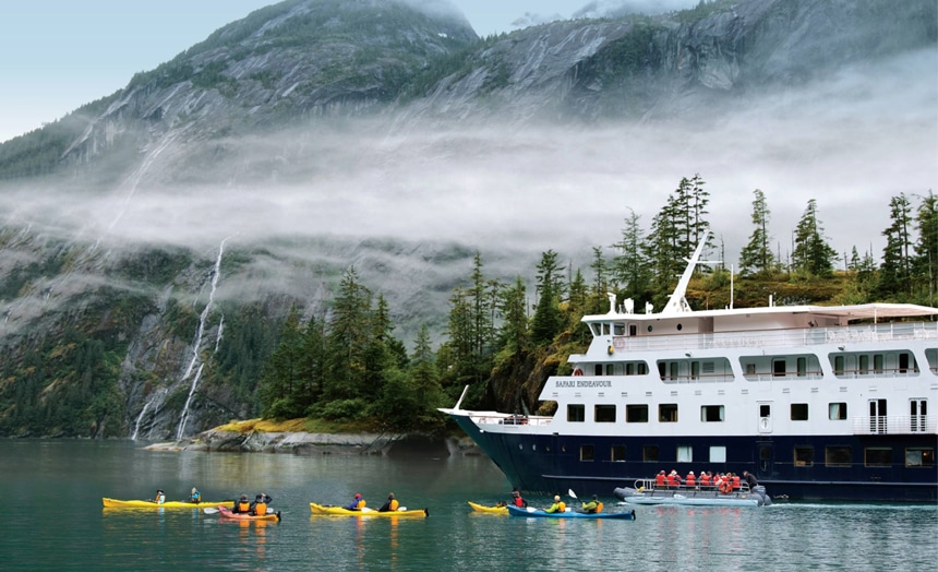 A misty fog morning, a white and blue ship floats as a group of kayakers paddle around it surrounded by a forested shoreline and mountain range,