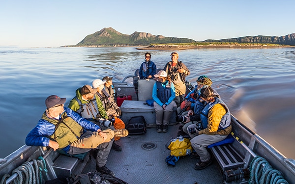 Nine travelers sit in a metal skiff on an Alaska small ship cruise sailing away from a mountainous shoreline.