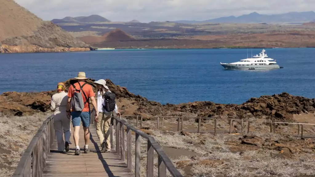 Aqua Mare Galapagos cruise travelers in shorts & t-shirts walk a wooden boardwalk over arid land as yacht sits offshore.