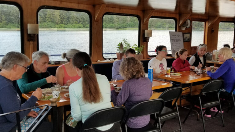 Travelers eating seated around a wooden dining table on the Snow Goose yacht in Alaska.