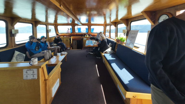 Salon on Snow Goose Alaska small ship with walls of windows, 2 dark blue benches filled with guests.