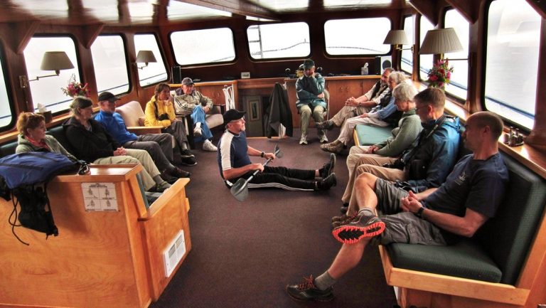 Salon on Snow Goose Alaska small ship with walls of windows, 2 dark green benches filled with guests & man sitting on floor.