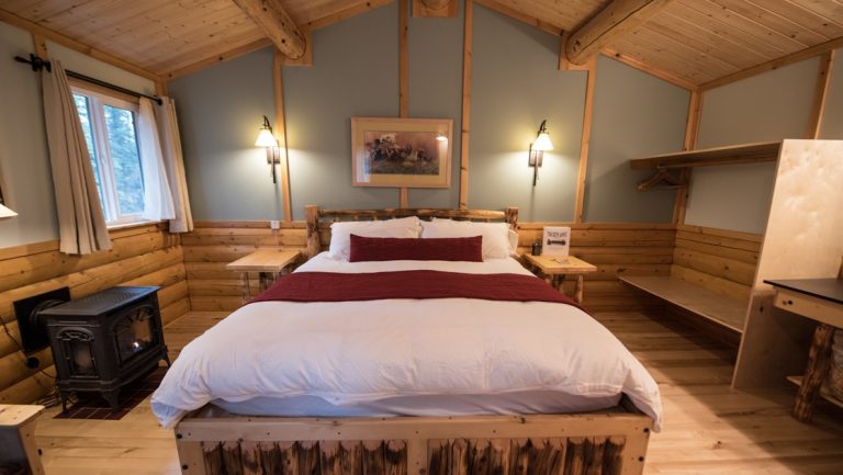 Tonglen Lake Denali lodge cabin interior with king bed, wood accents, bright windows, lamps & small cast iron stove.