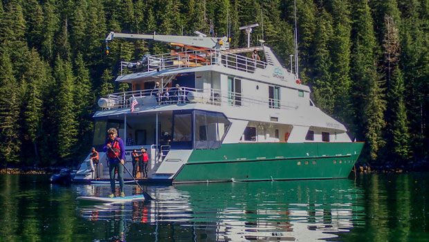 Small yacht with green hull & 2 white upper decks sits in calm water as woman paddleboards nearby on San Juan Island cruises.