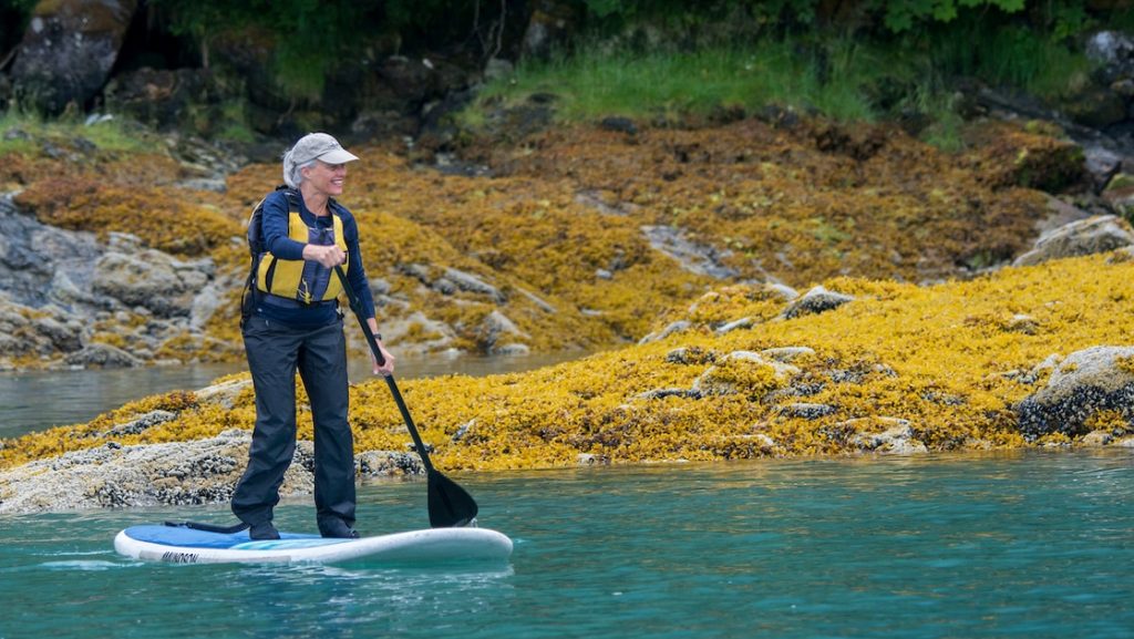 A solo Alaska traveler seen paddling on a white paddleboard with bright yellow moss on the shoreline behind her