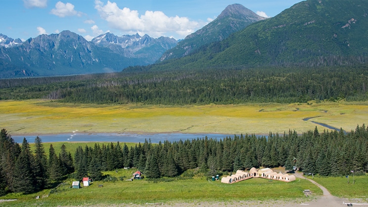 Aerial view of small camp with modern beige hoop tents set amongst forest, river, open grassland & large mountains in Alaska.