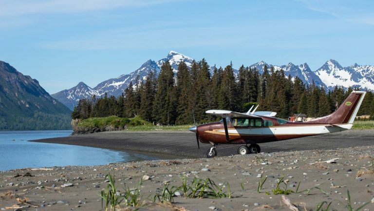 Small bush plane with brown, green & white paint sits on a remote, empty beach on a sunny day in Alaska.