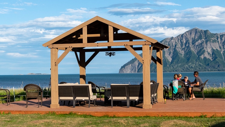 Bear Camp Alaska guests sit beside large wooden bandstand at the water's edge on a sunny day.