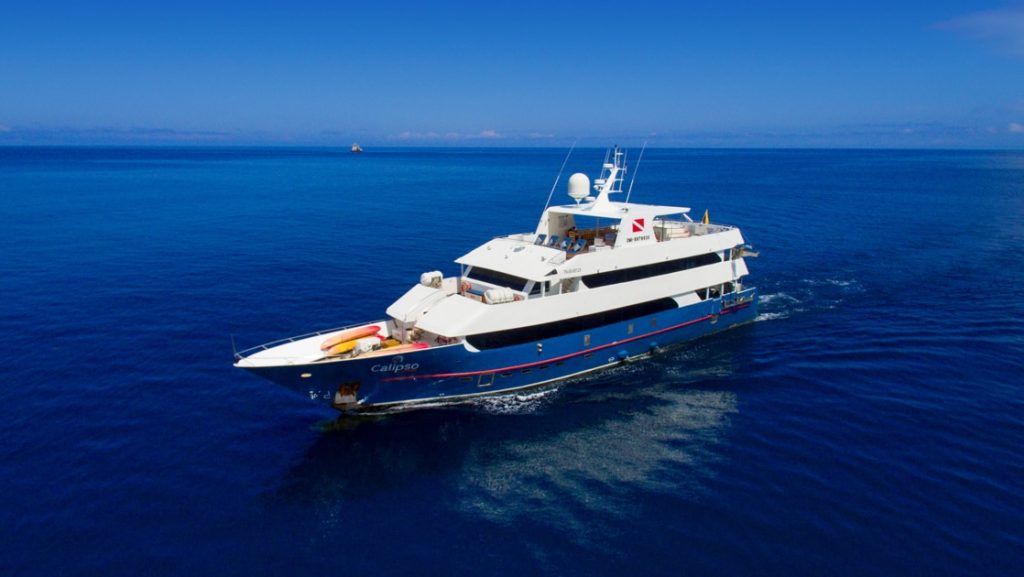 Aerial view of Calipso yacht in Galapagos with dark blue hull & white upper decks, cruising through calm blue water on a sunny day.