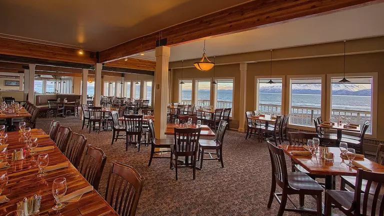 Land's End Resort Homer Alaska dining room with long tables set for dinner service, dark wood furniture & ocean views.