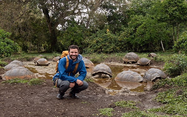 A man in a blue jacket and backpack squats in front of a small pond full of Galapagos giant tortoises