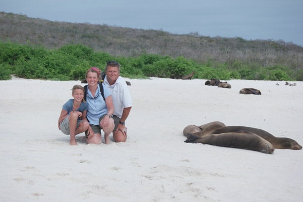 A father, mother and young teen daughter pose on a white-sand beach next to sleeping seals on their Galapagos Island vacation.