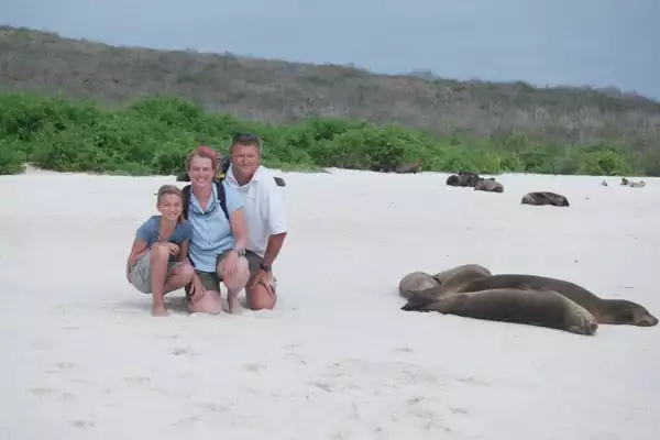 A father, mother and young teen daughter pose on a white-sand beach next to sleeping seals on their Galapagos Island vacation.