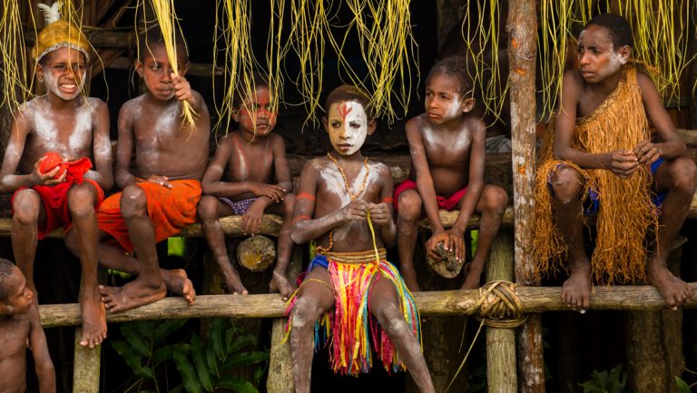 Boys with painted faces & grass skirts sit in a line for a greeting ceremony of tribal culture in Asmat, New Guinea.